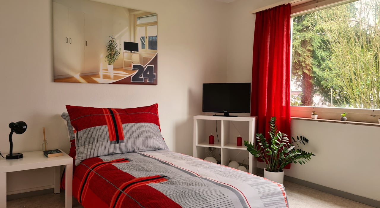 Spacious bedroom with large windows, red and gray patterned bedding, white furniture including a dresser and shelving unit, and a TV mounted on the wall.