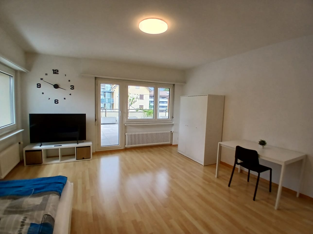 The image shows a studio apartment with a large open floor plan. The room has hardwood floors, white walls, and large windows that provide natural light. There is a TV stand with a flat-screen TV, a wardrobe, and a desk with a chair. The apartment appears