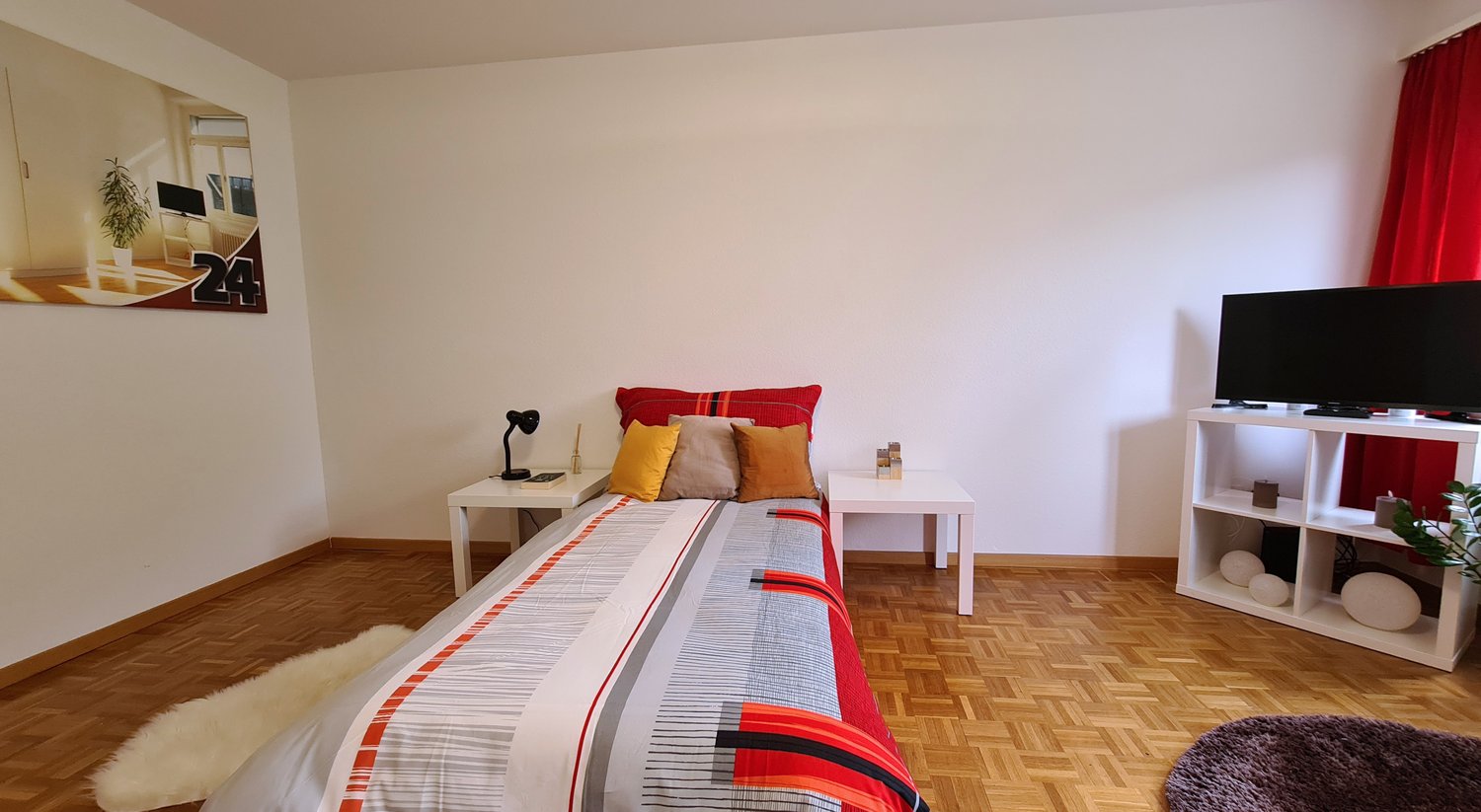 Spacious bedroom with hardwood flooring, white walls, a large bed with colorful bedding, side tables, a lamp, and a TV stand. The room also has a window, suggesting the presence of a balcony or terrace.