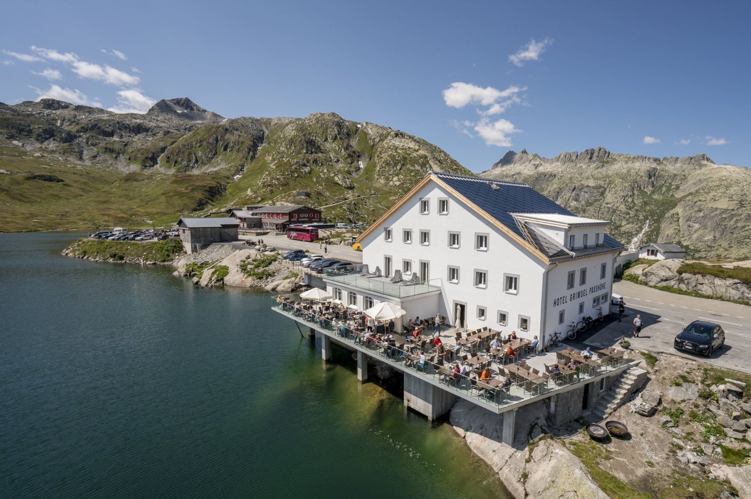 A white hotel building with a terrace overlooking a scenic mountain lake surrounded by rocky peaks. There are people dining and relaxing on the terrace. The hotel appears to be situated on a cliff or elevated platform with a parking area below.