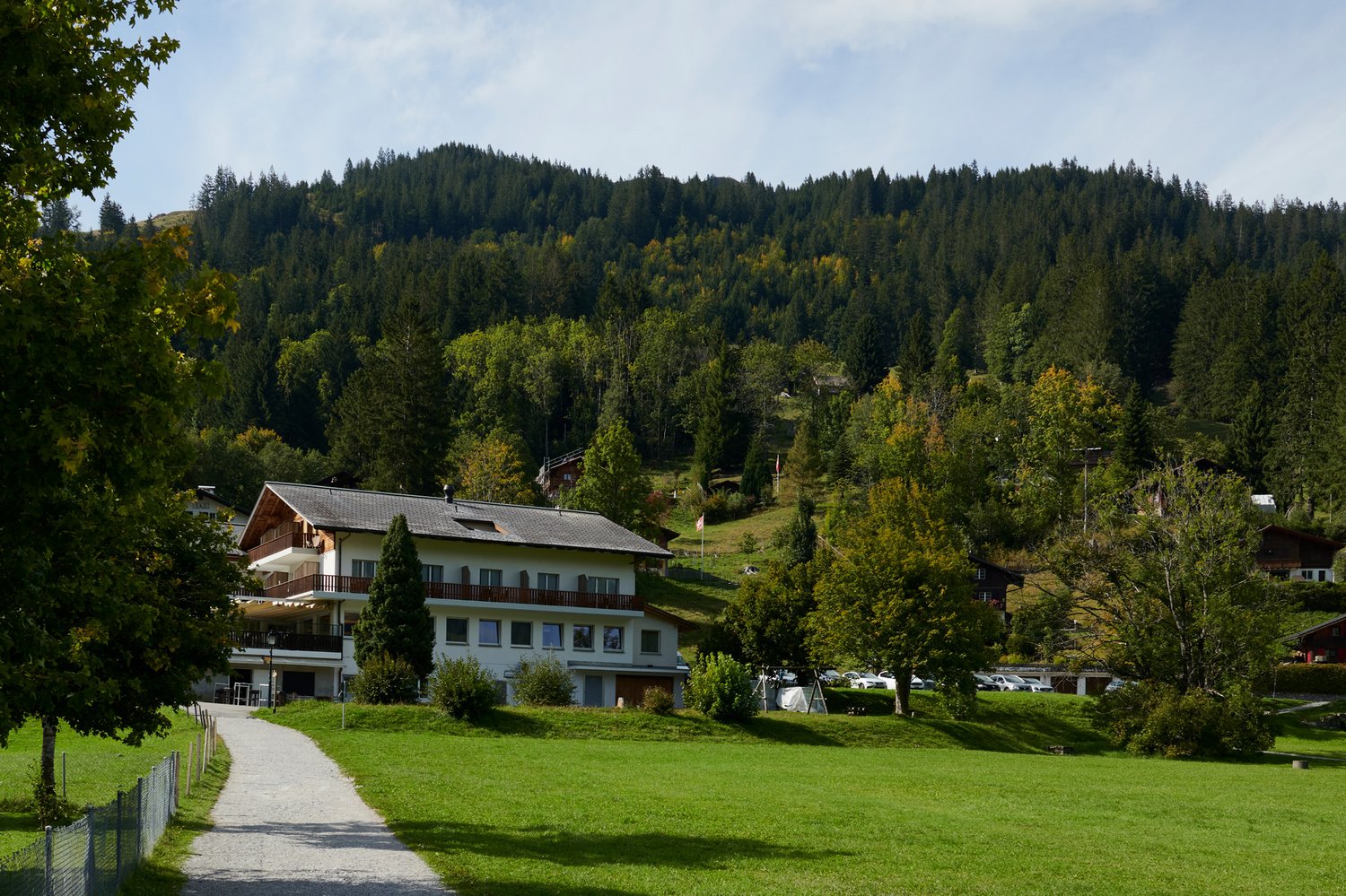 Mountain-side building with a balcony, surrounded by green meadow and lush trees, offering a breathtaking mountain view