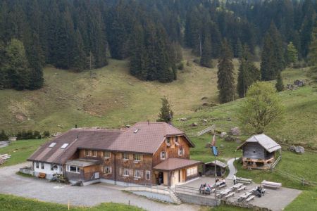 Two-story wooden house with a brown roof, nestled in a green valley, surrounded by mountains, large windows, and a small outbuilding. In front of the house, there is a paved area with a few tables and benches for outdoor seating.