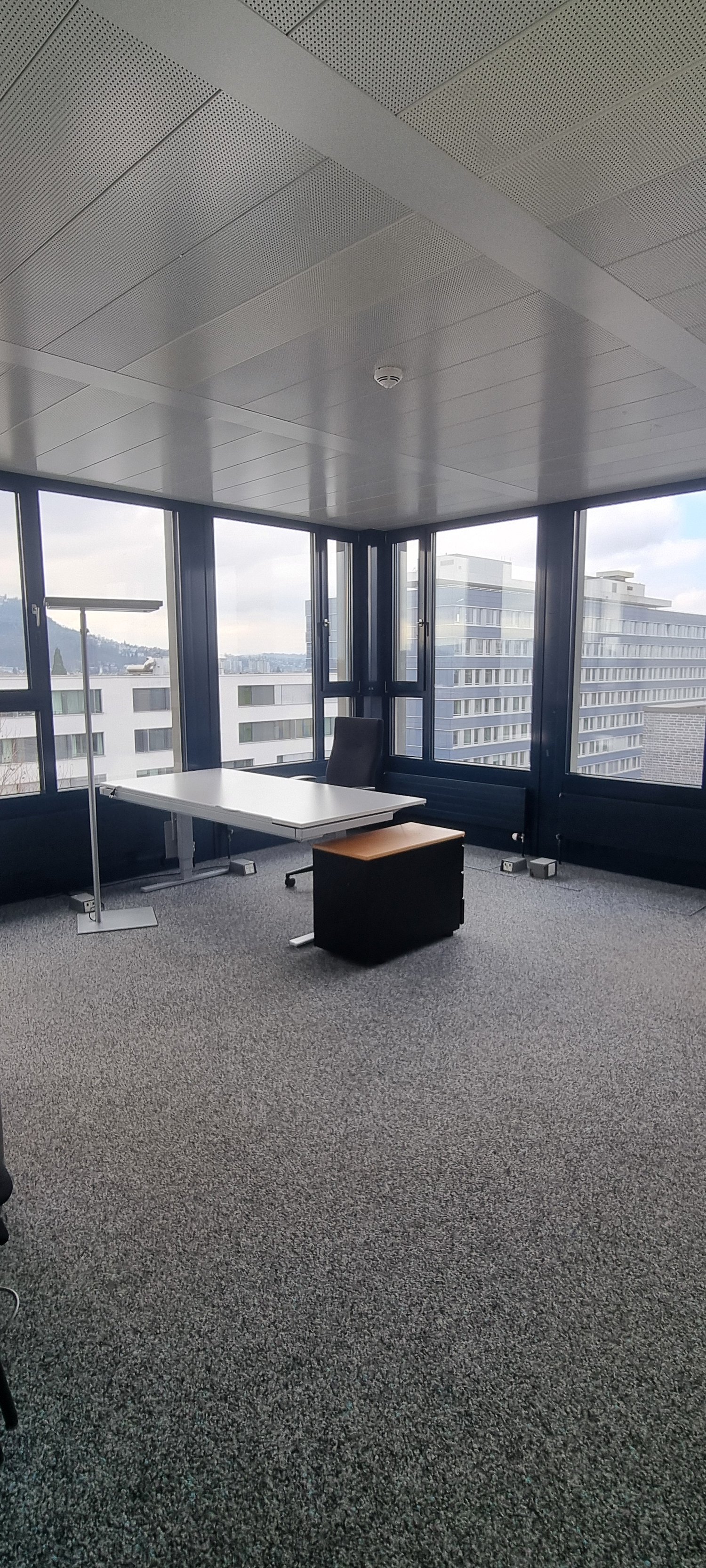This image shows a modern office space with large windows providing a view of the surrounding cityscape. The room has a minimalist design with a white table, a black office chair, and a gray carpeted floor. The ceiling features a grid-like pattern of pane