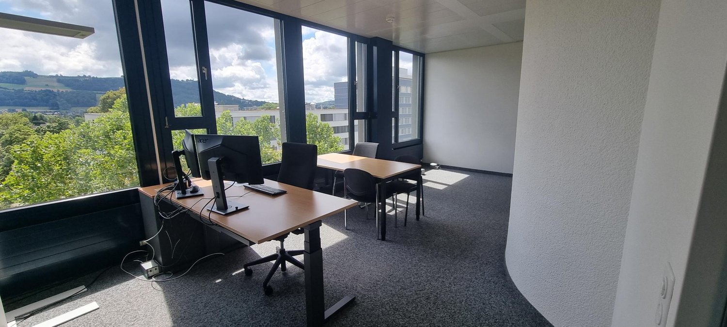 Spacious office with large windows offering a scenic view of the surrounding landscape, including hills and greenery. The office is equipped with desks, chairs, and computer equipment, providing a functional workspace.
