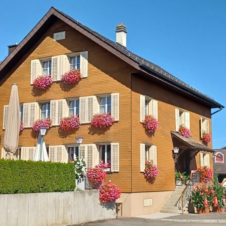 2 story house, brown wooden exterior, white shutters, pink flowers in window boxes, garden with hedges and plants, awning over entrance