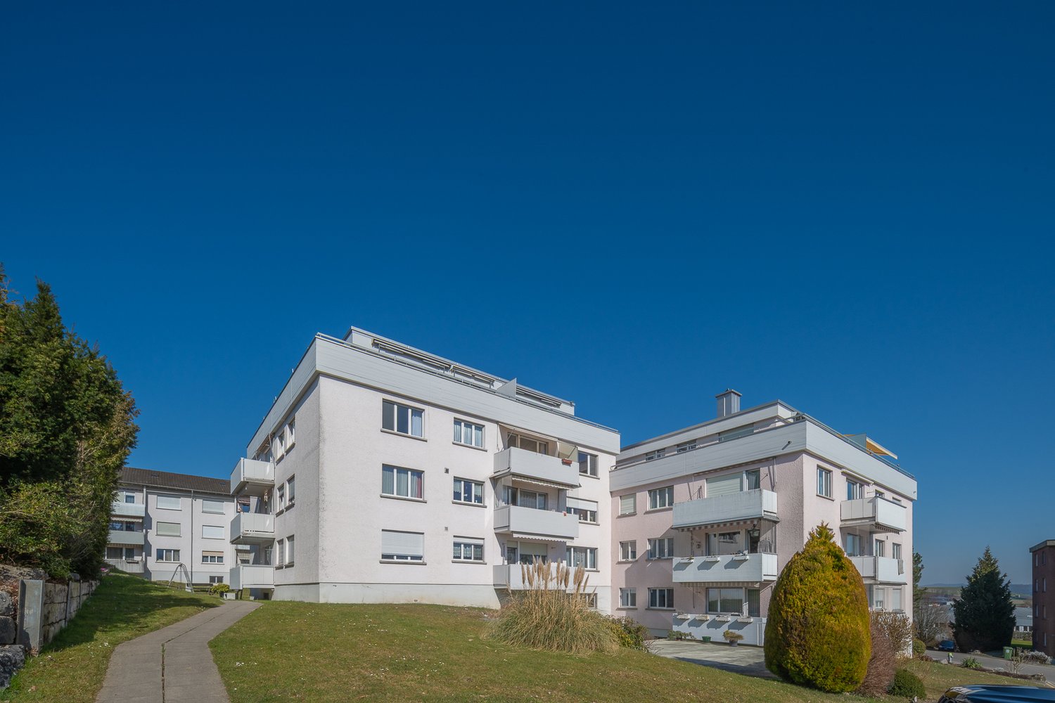 Modern apartment building, white exterior, multiple balconies, landscaped garden, driveway