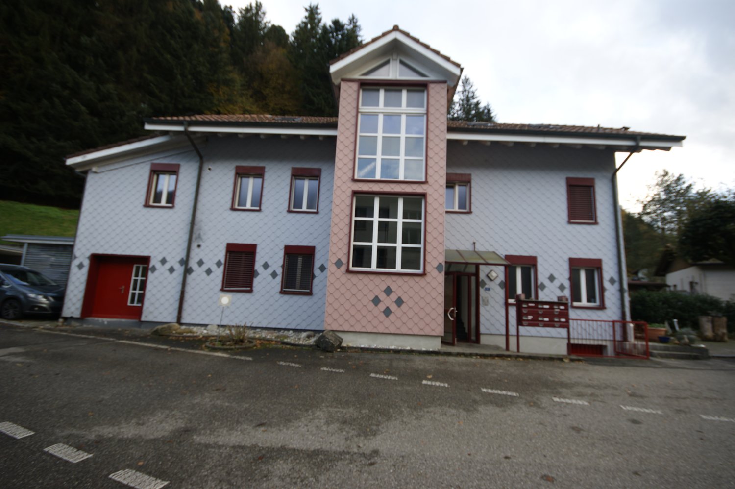 Two-story building, white and pink facade, multiple windows, red entrance door, brown roof, located on a road