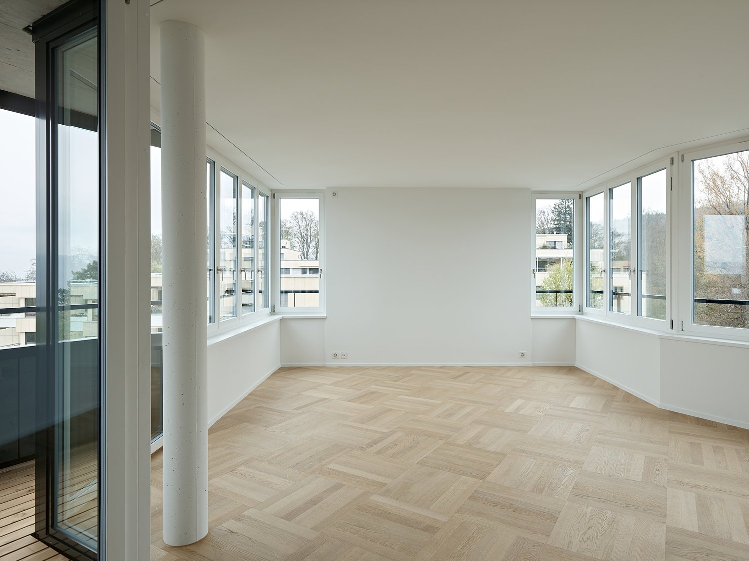 Large room with wooden parquet flooring, white walls, glass windows, central white pillar, open balcony with balcony railings