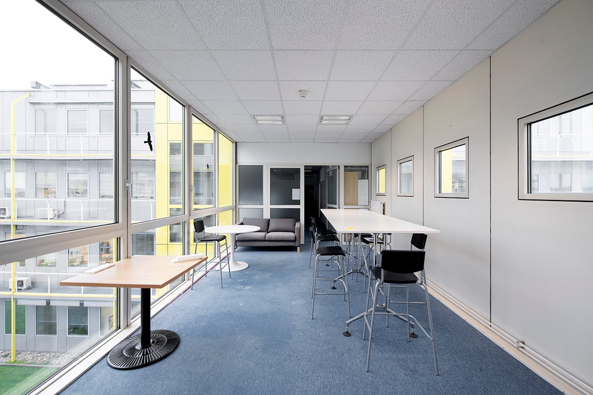 Large open office space with floor-to-ceiling windows, gray carpeted floors, white walls, and modern furniture including a sofa, tables, and chairs. The space appears to be designed for a professional work environment.