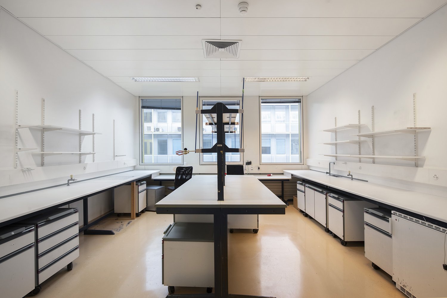 The image shows a modern, well-lit office space with a large central table, shelving units along the walls, and various workstations. The room has large windows providing natural light, and the overall design appears to be minimalist and functional.