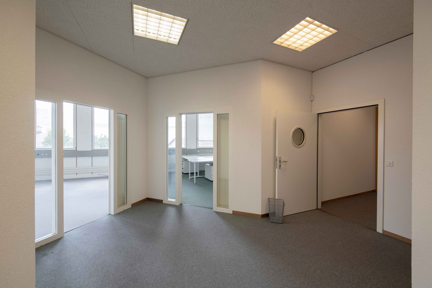 Empty room with white walls, gray carpeted floor, glass doors and windows, round window in door, trash can, desk with chair in the background, ceiling lights