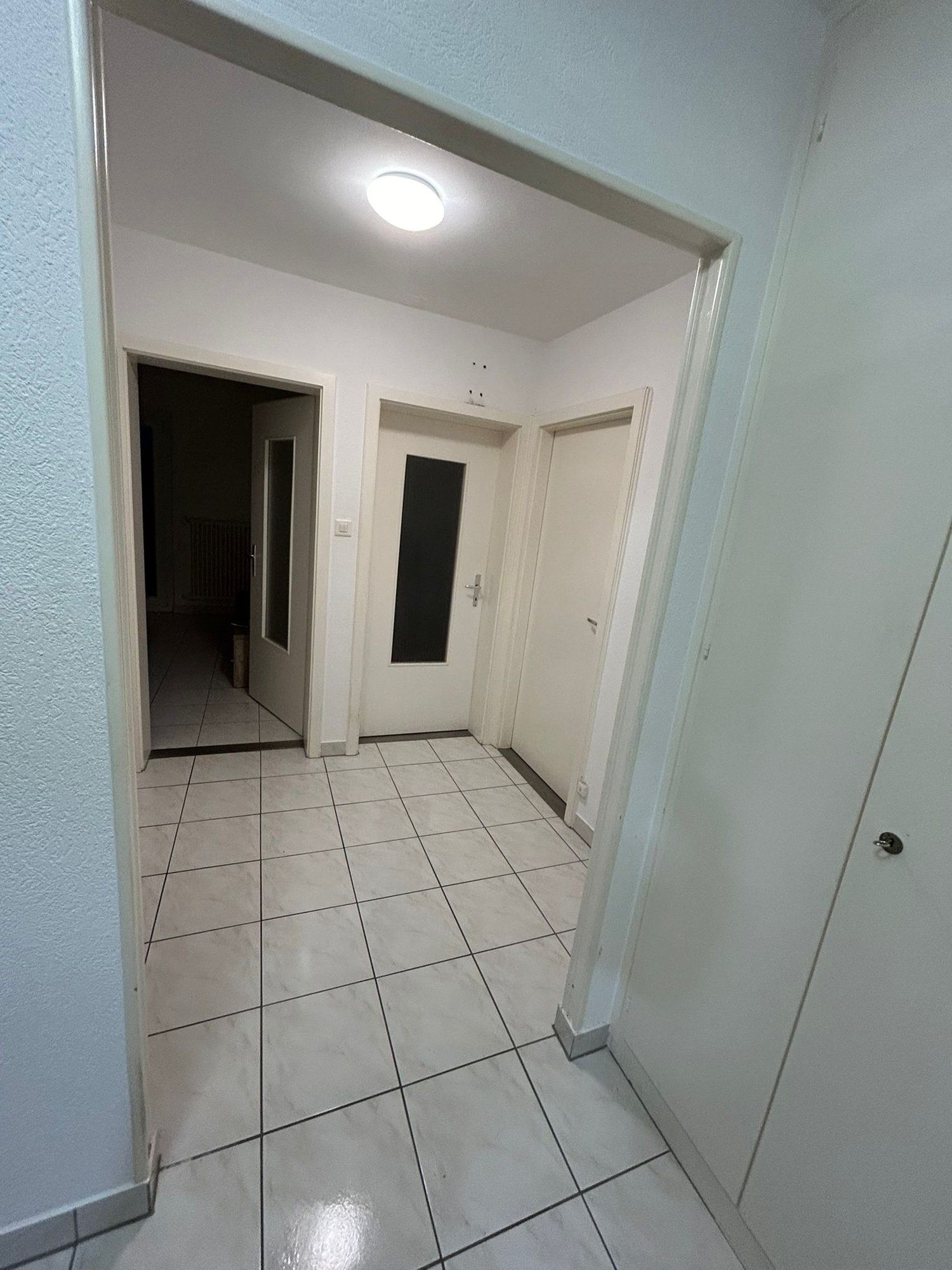 Empty hallway with white tiles, white walls, and white doors. A ceiling light is on.