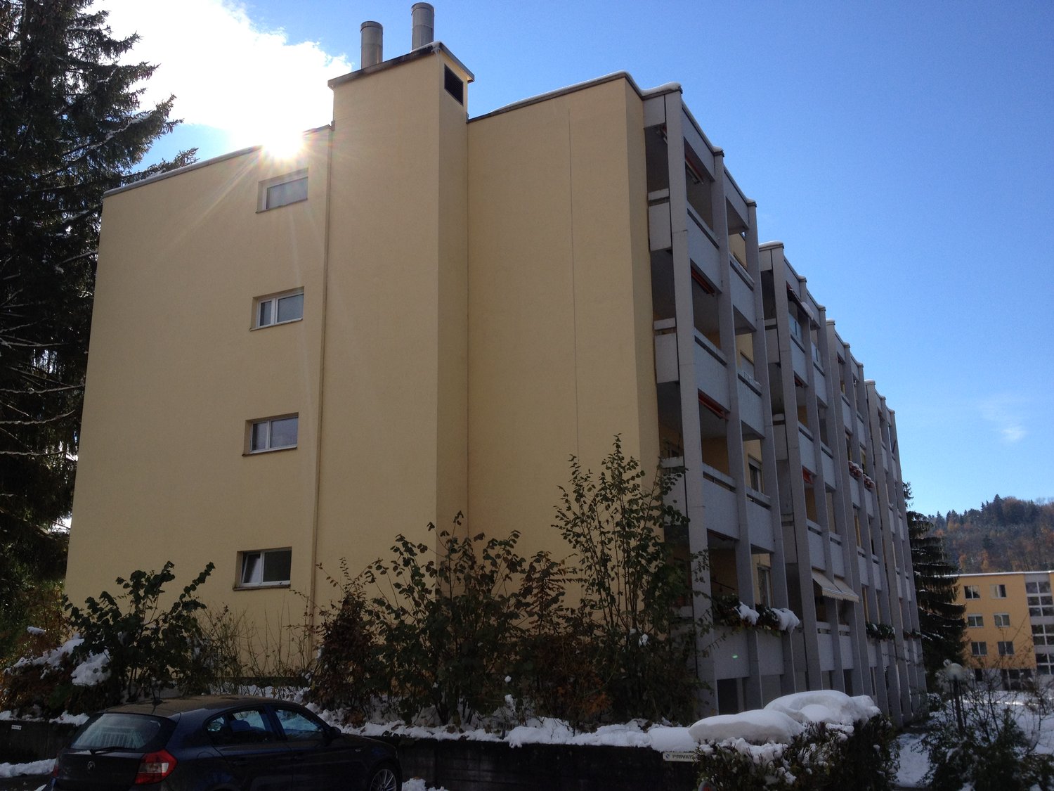Multi-story building, yellow exterior, multiple windows, balconies, snow on the ground, car parked in front