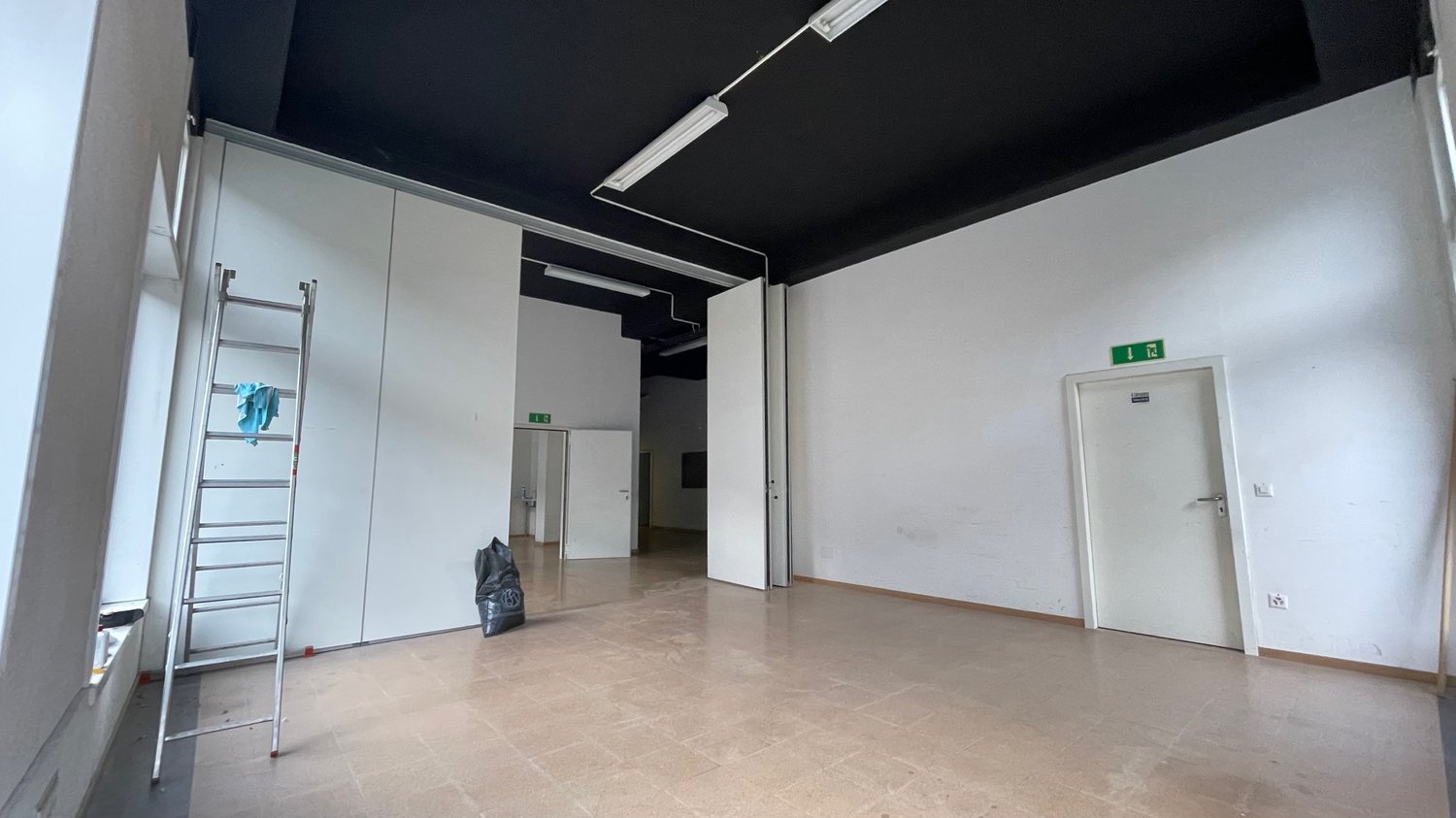 Empty room, black ceiling, white walls, tiled floor, open doors, metal ladder, trash bin