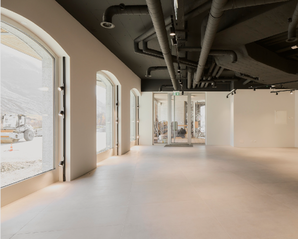 Large open commercial space with high ceilings, exposed pipes, and large windows overlooking a parking area
