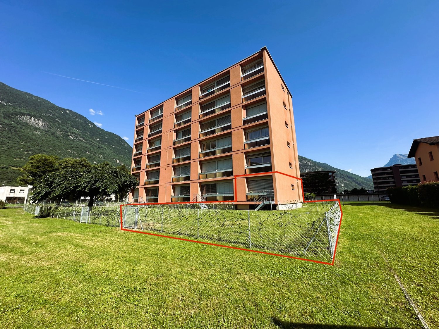 large orange building, many balconies, large red square outlined, tall green trees, grassy ground, mountains in background