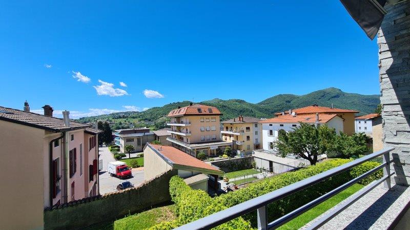 View from a balcony with a railing, overlooking a street with buildings, mountains, a car, and a tree.