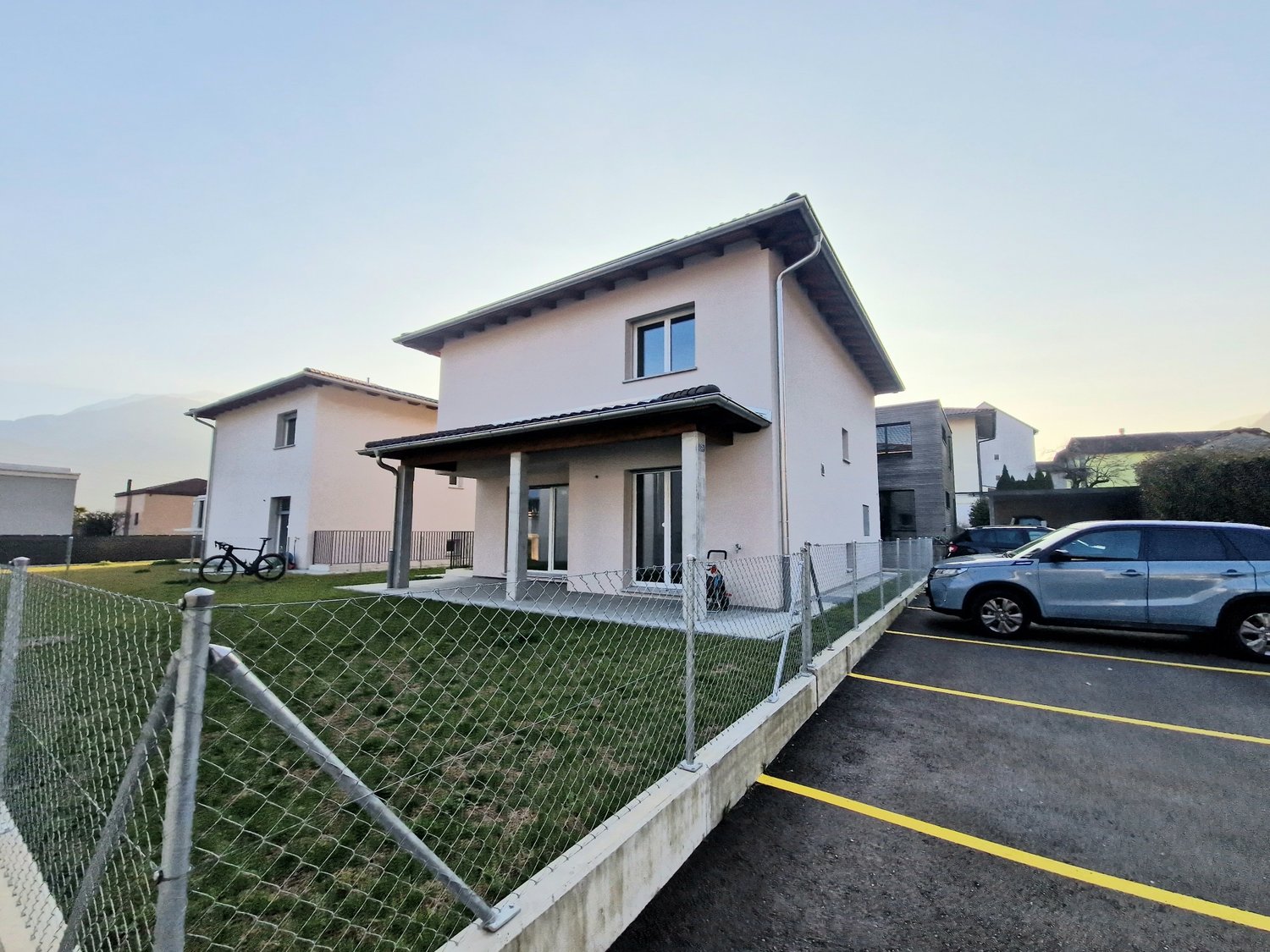 Modern two-story house, white exterior, large windows, bicycle parked in front, fenced area, SUV parked in parking lot