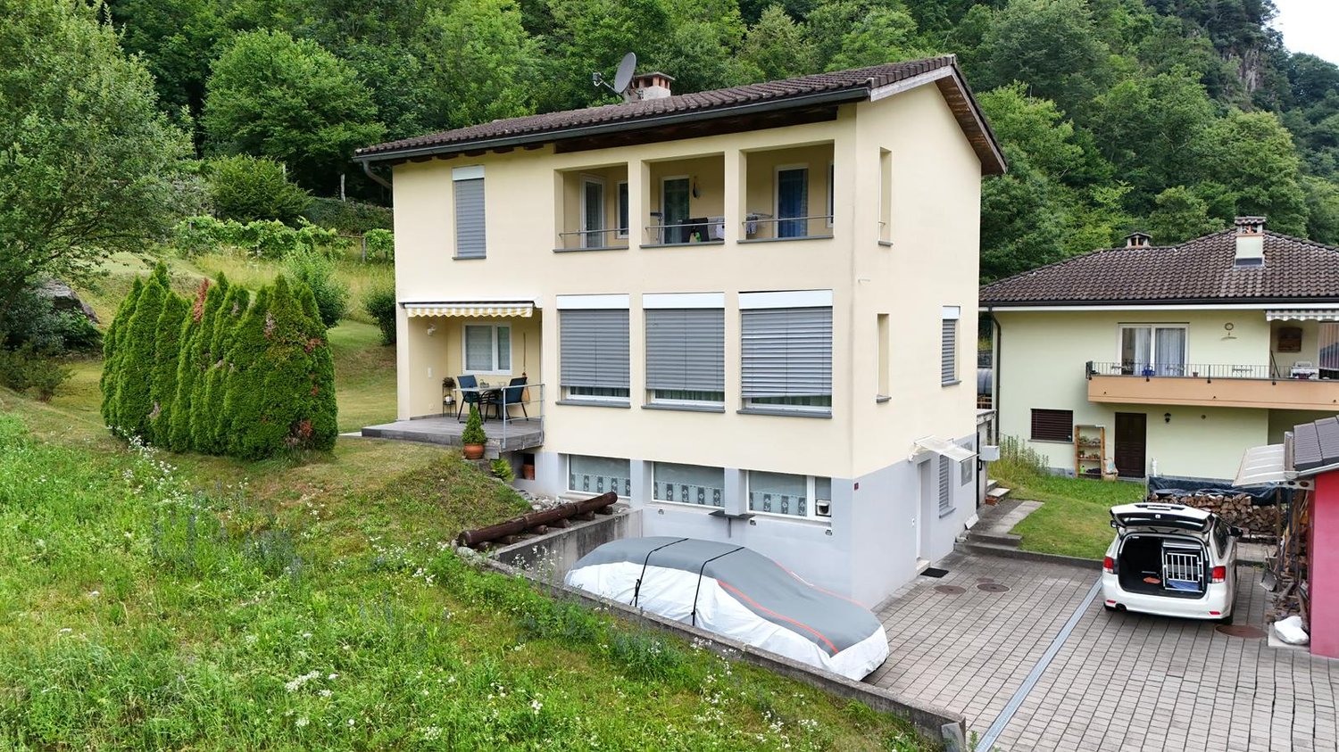 Two-story house with a yellow exterior, balconies, and a paved driveway with a parked car. The house is surrounded by lush greenery and trees.