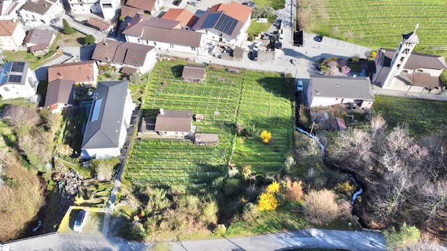 Aerial view of a residential area with several houses, a church, and a small stream. The houses are surrounded by green fields and trees. Some of the houses have solar panels on their roofs.