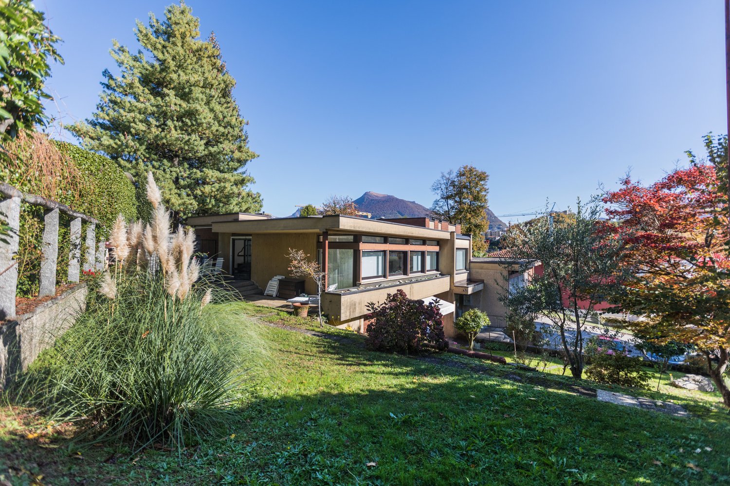 modern-style house, yellow walls, 3-story, large windows, outdoor seating, lawn, trees, mountains in background
