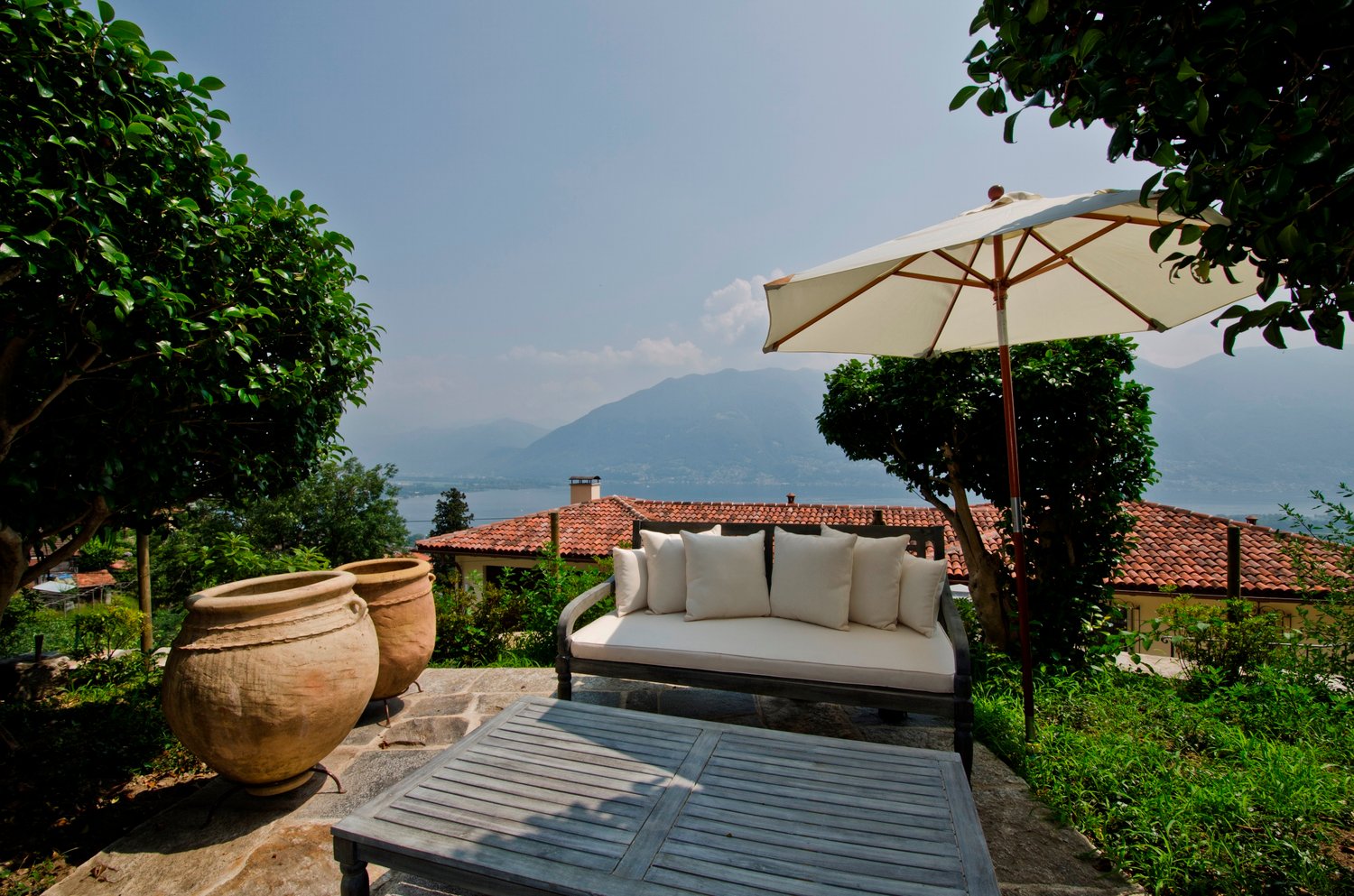 The image shows an outdoor patio area with a comfortable sofa, a large umbrella, and two large terracotta pots. The patio overlooks a scenic landscape with mountains in the background, suggesting a beautiful view. The patio is surrounded by lush greenery,