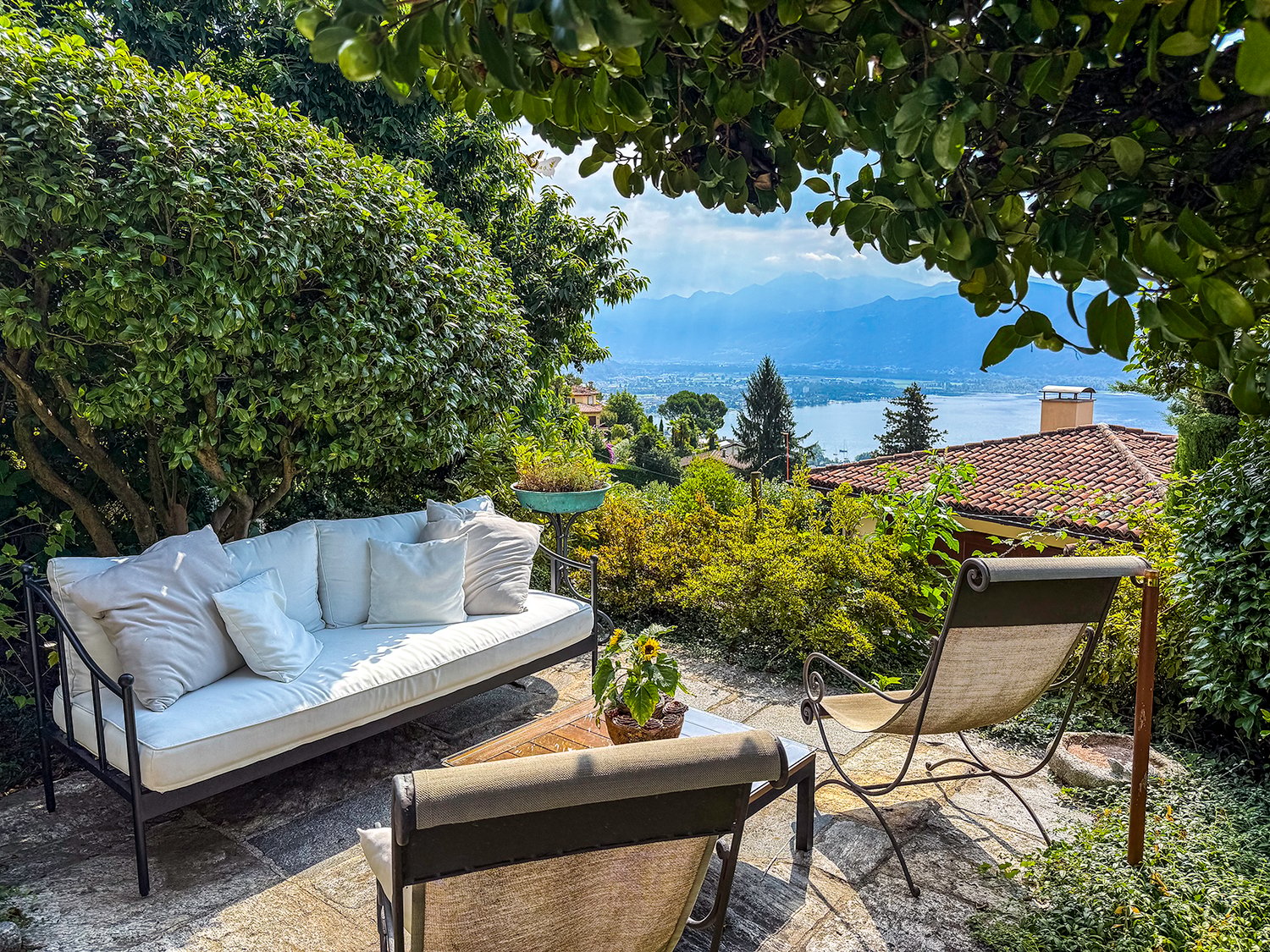 Cozy outdoor patio with a comfortable sofa, chairs, and a scenic view of the lake and mountains in the background, surrounded by lush greenery and trees
