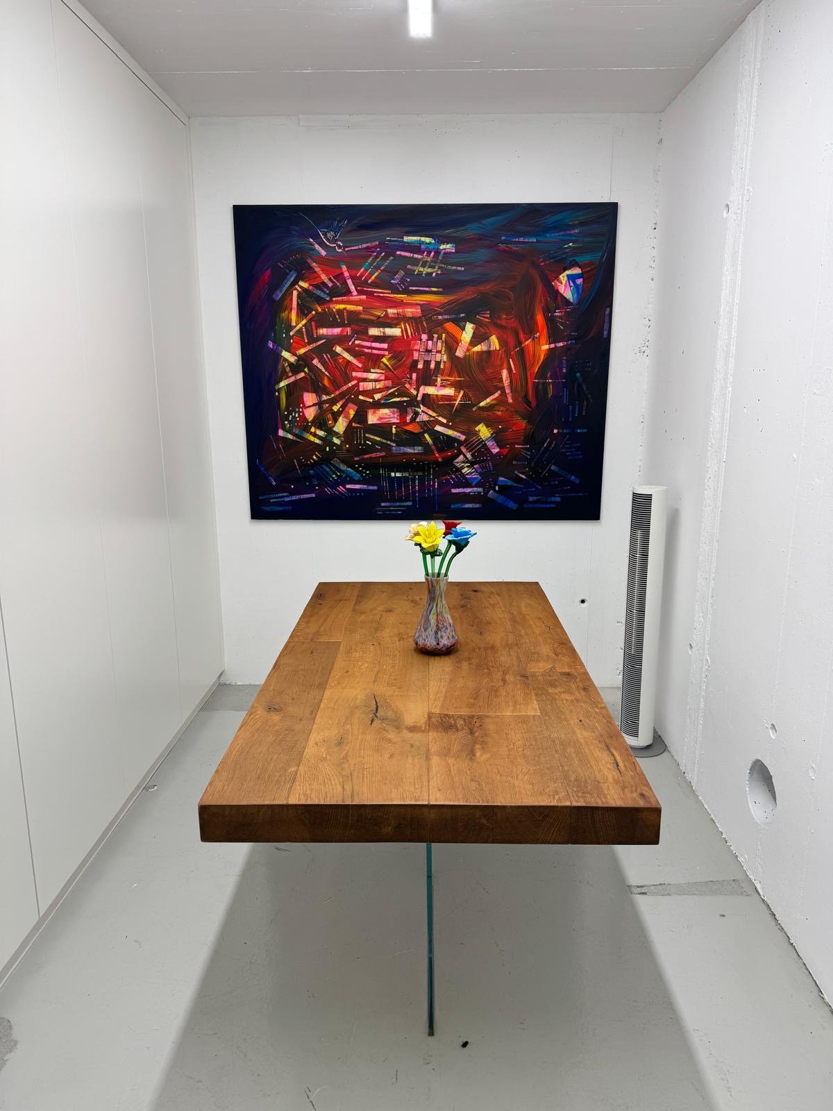 Dining area with a wooden table, colorful flowers in a vase, large painting, and white walls
