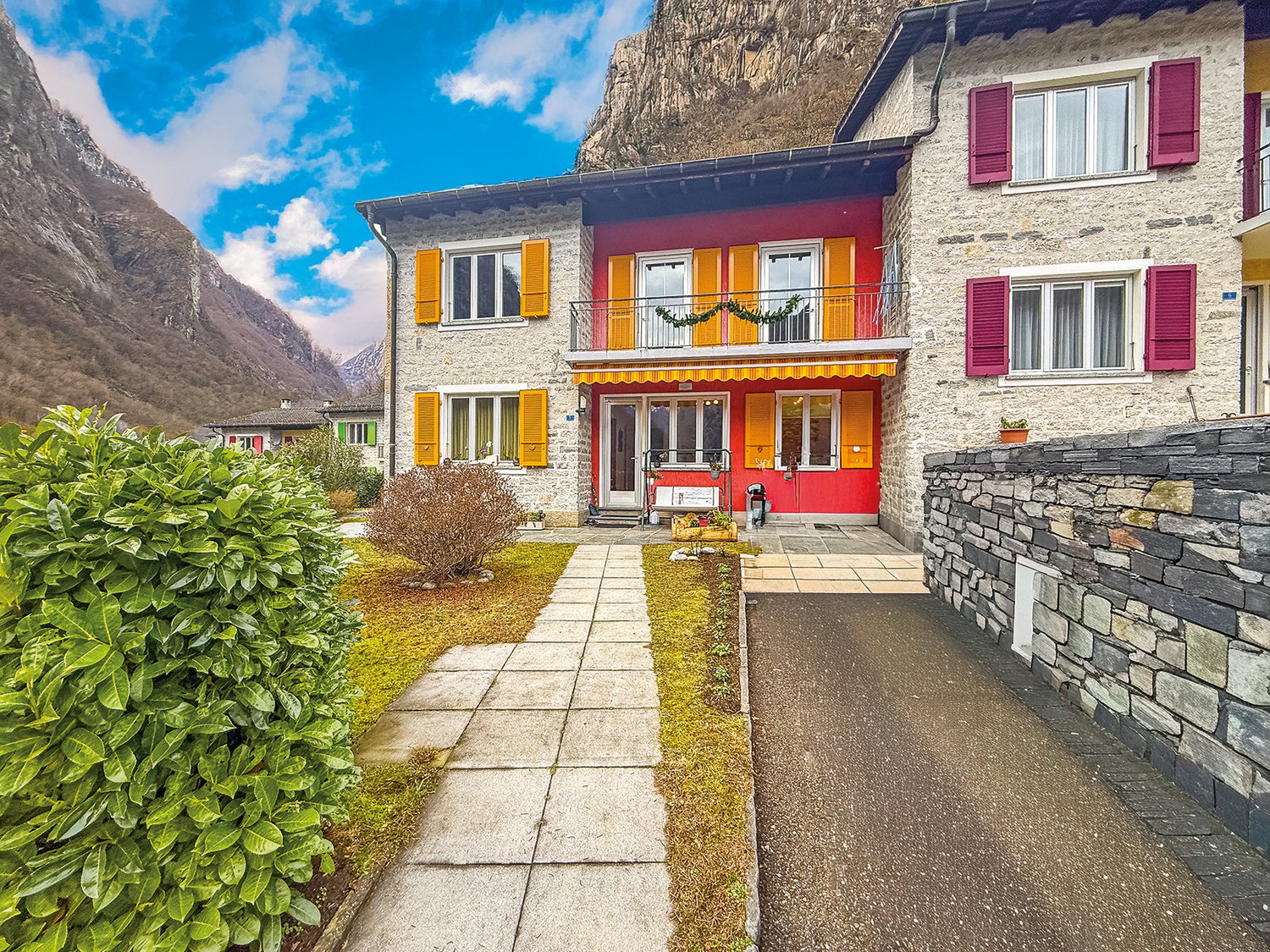 Mountain villa with red wall, white shutters, yellow shutters, white door, entrance paved walk, small bushes, stones on the side, pathway, small garden area