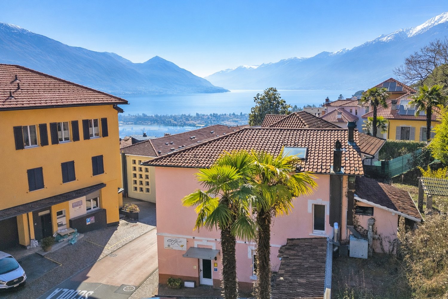 Aerial view of buildings with red roofs and yellow facades, overlooking a lake and mountains, surrounded by greenery.