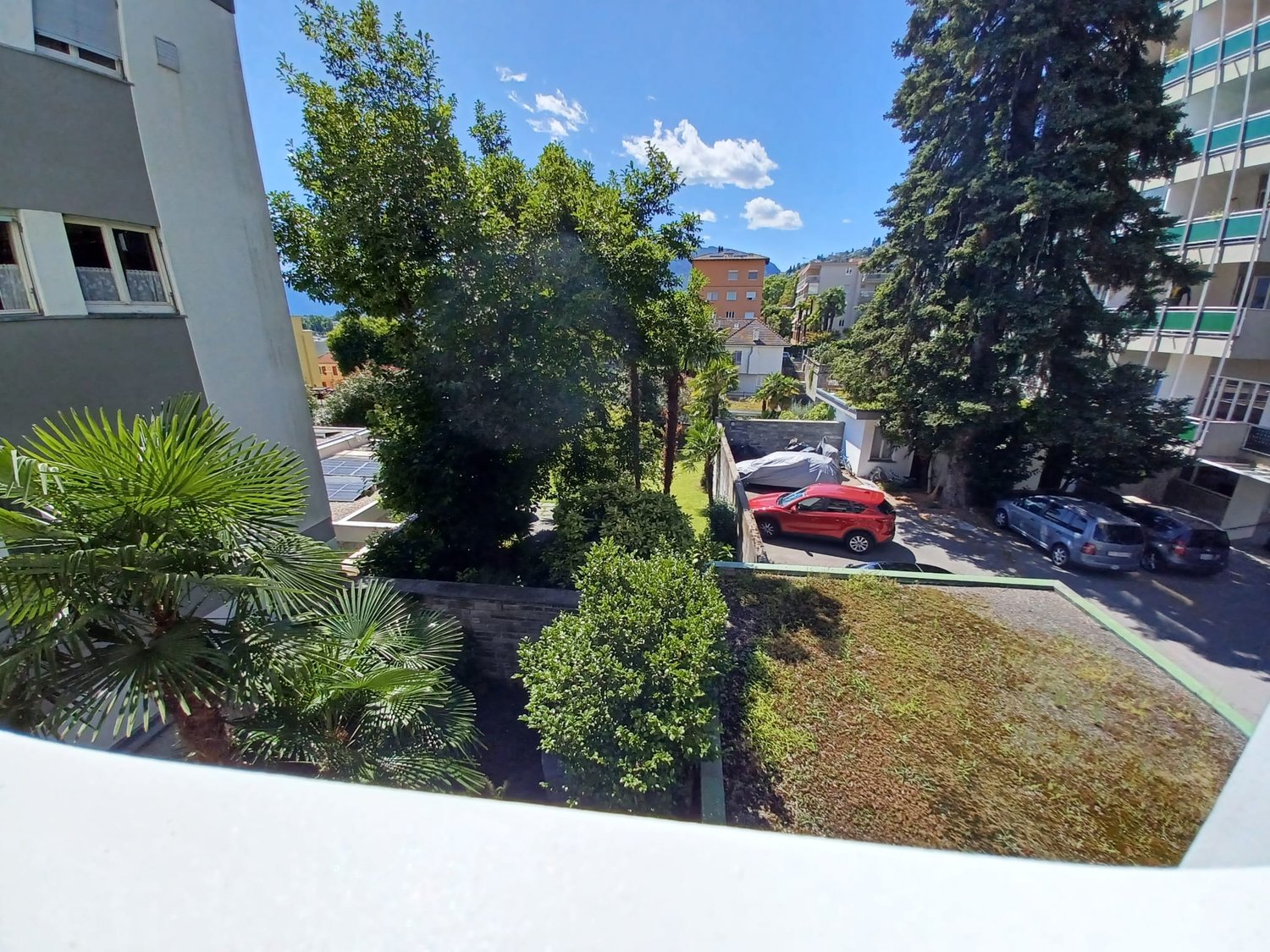 View of a parking area with several cars parked, surrounded by trees and bushes, with a building in the background.