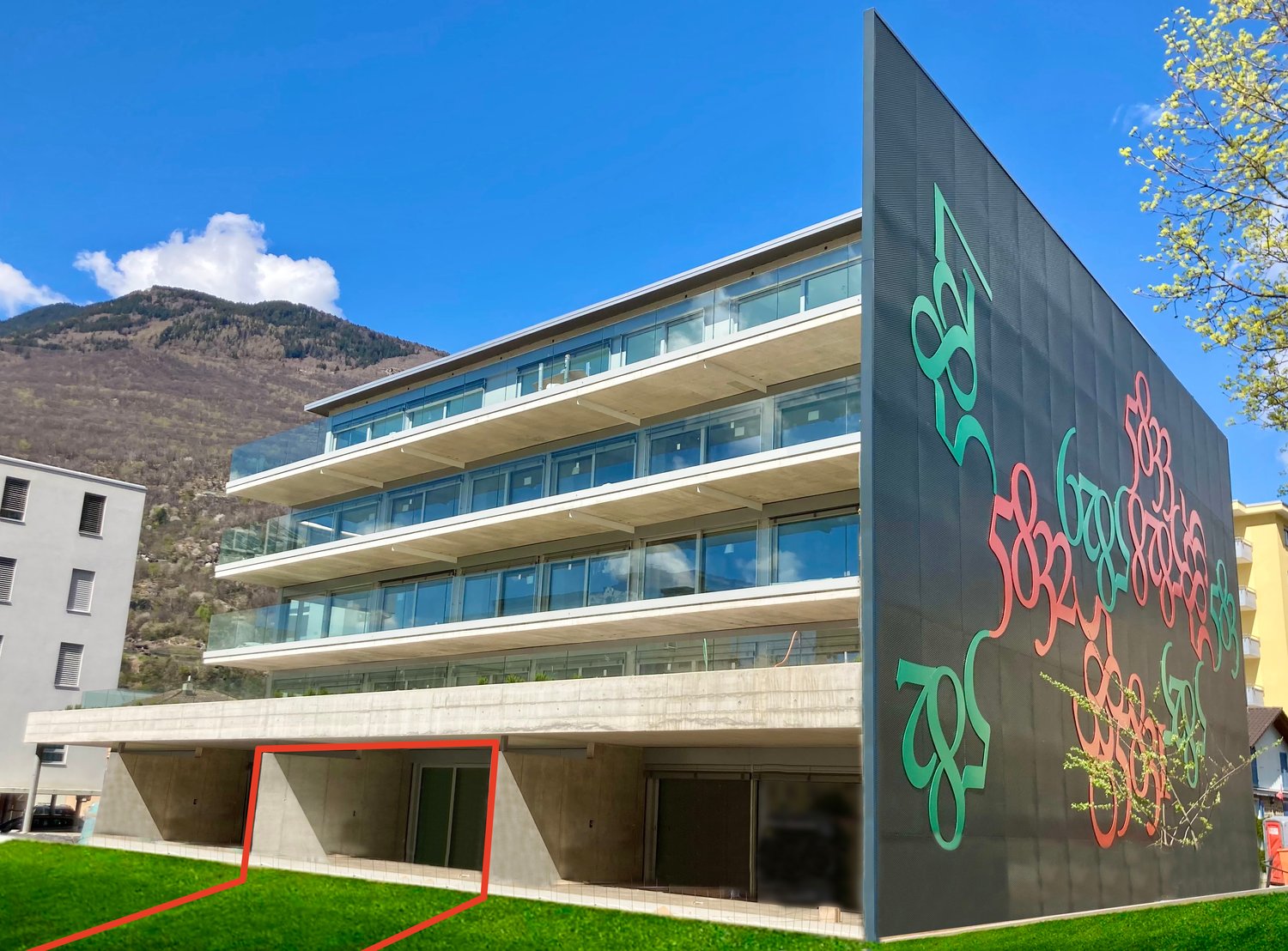 Modern building with glass windows, concrete structure, graffiti on the wall, green lawn, mountains in the background