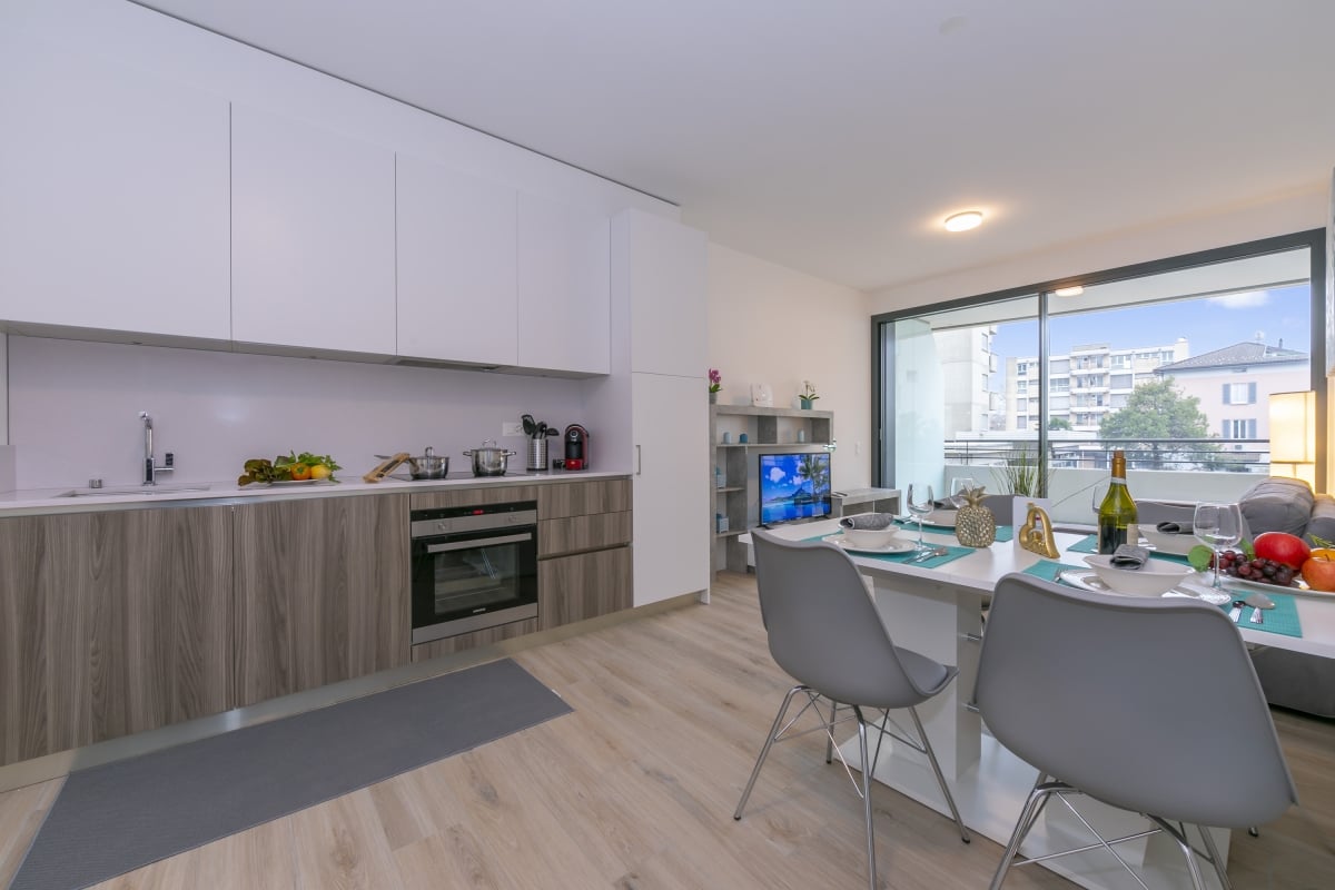modern kitchen, wooden cabinets, countertop, sink, faucet, electric oven, balcony with view, dining table with chairs, TV, shelf, bowl, bottle, wine glasses, grapes, apples, pineapple, lamp, window