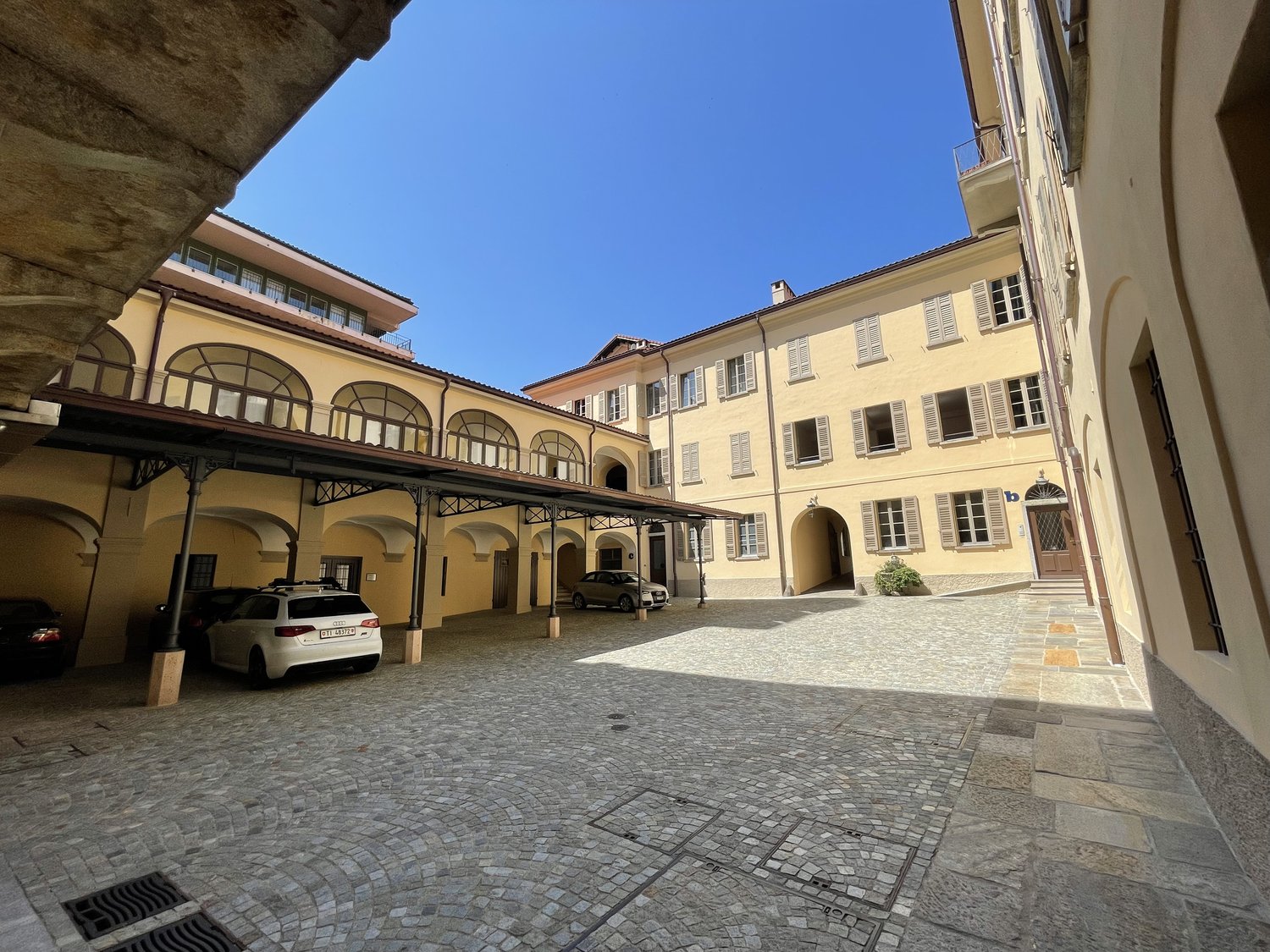 Cobblestone courtyard with cars parked under a covered area, yellow building with arched windows and doors, balconies, and a small plant in front of the entrance.