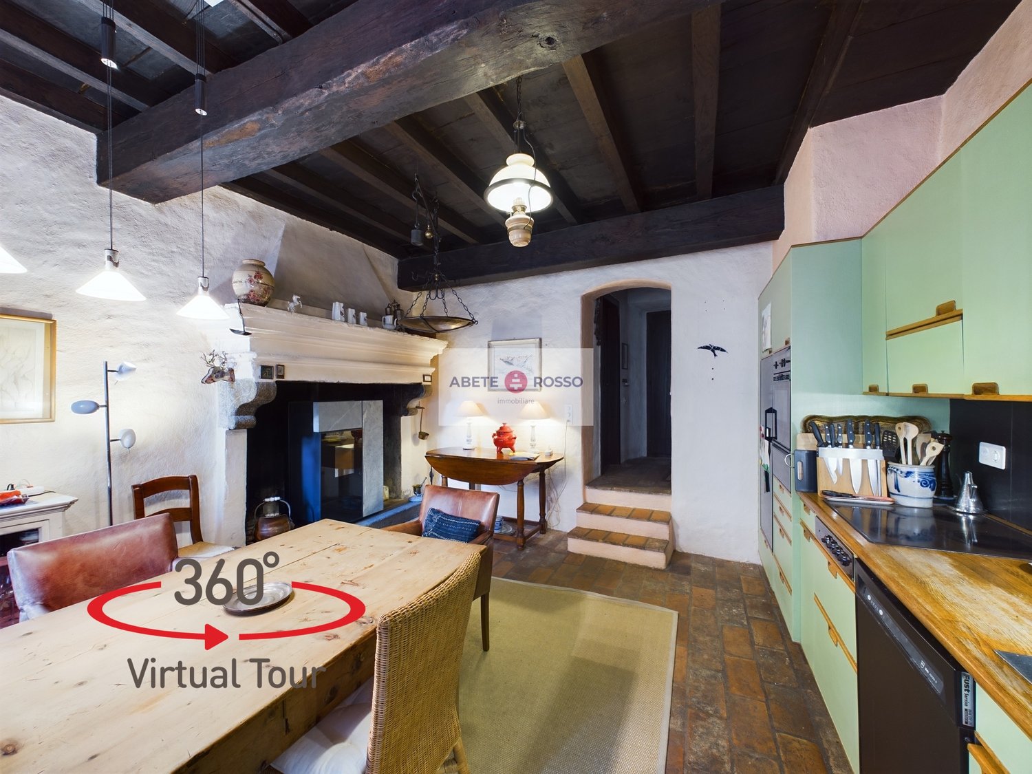 Rustic kitchen with exposed wooden beams, stone walls, and a fireplace. The kitchen features a wooden dining table, wicker chairs, and various kitchen appliances and utensils. There is also a 360 degree virtual tour sign visible.