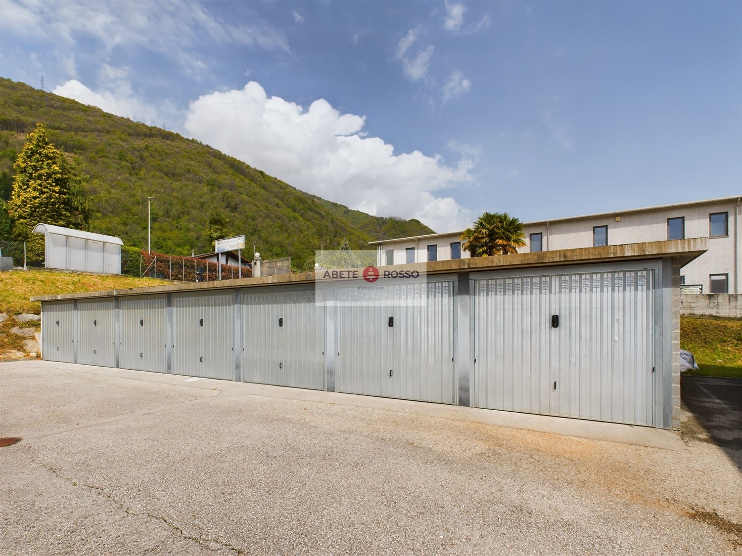 Garage building with multiple storage units, concrete floor, sign reading 'ABETE ROSSO Immobiliare', surrounded by greenery and a mountain in the background