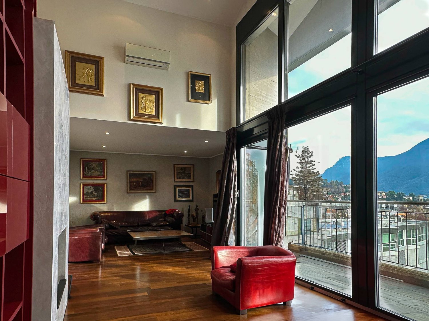 Spacious living room with large windows offering a scenic view, hardwood flooring, leather sofa and armchair, framed artwork on the walls, and air conditioning unit