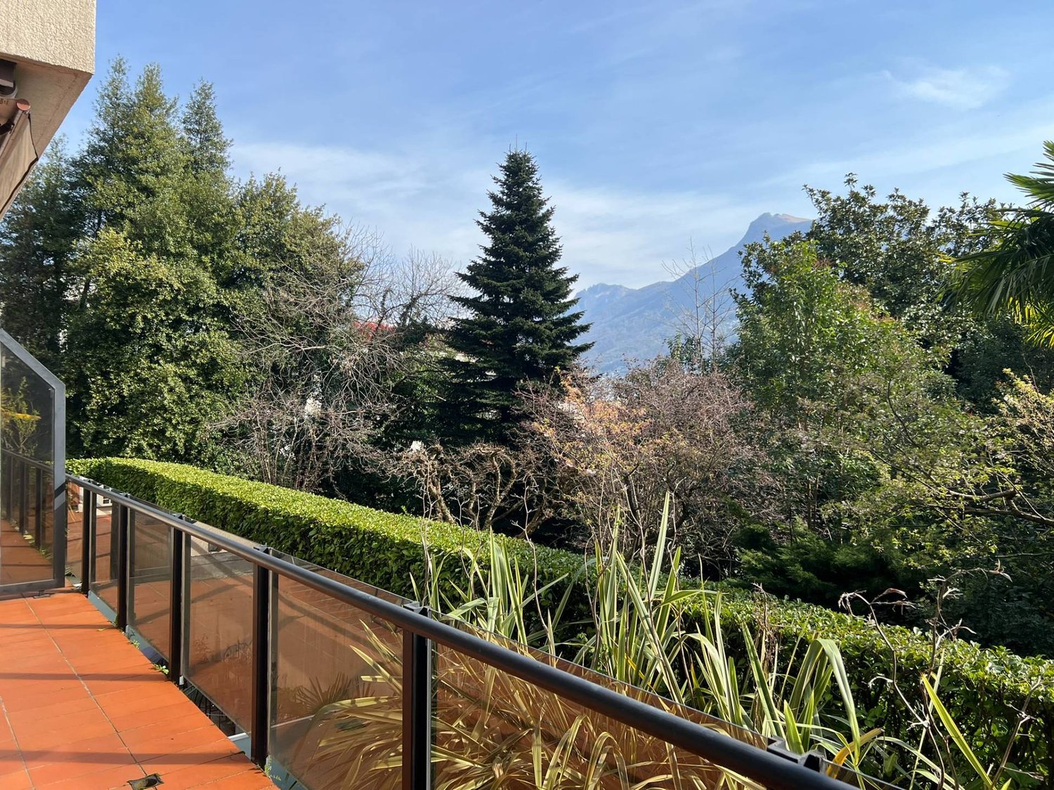 Balcony with glass railings, tiled floor, hedges, trees, mountain view