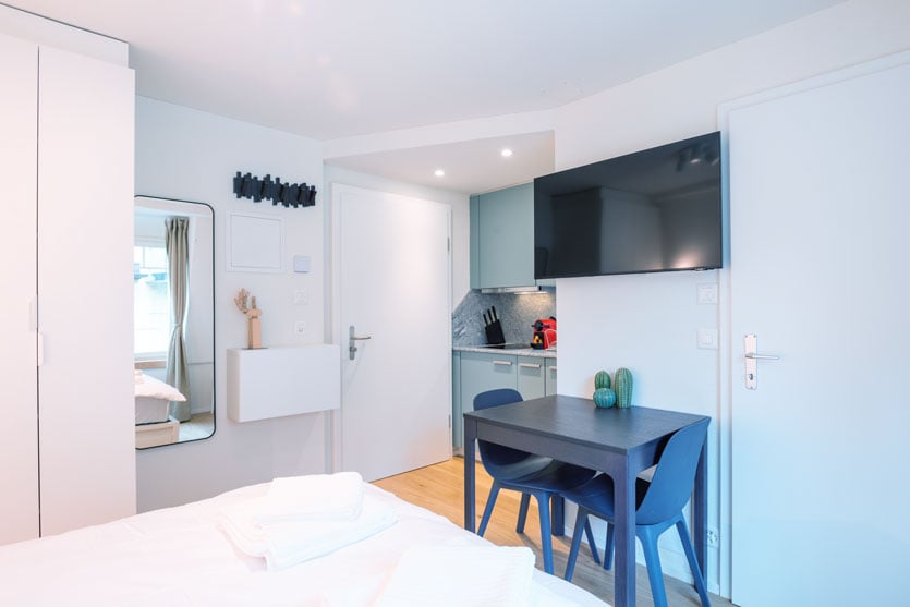 Compact studio apartment with open-plan layout, white walls, hardwood floors, kitchenette with dishwasher and washing machine, dining table, and a large TV mounted on the wall
