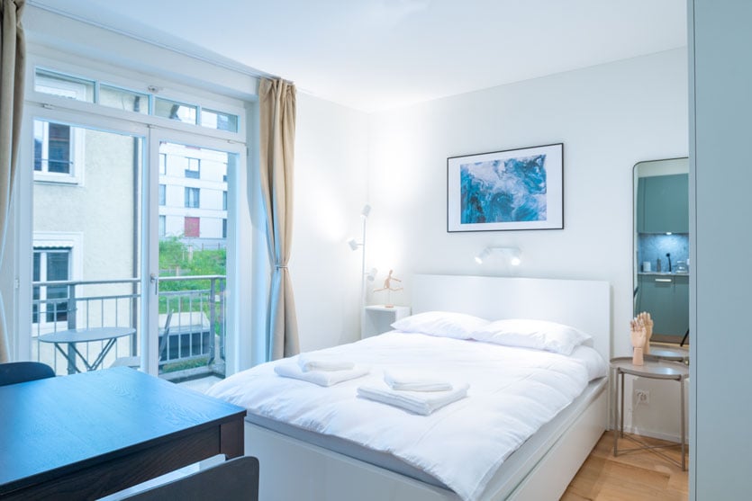 Bright and airy bedroom with large windows, white walls, and a balcony. The room features a double bed, a bedside table, and a framed artwork on the wall.