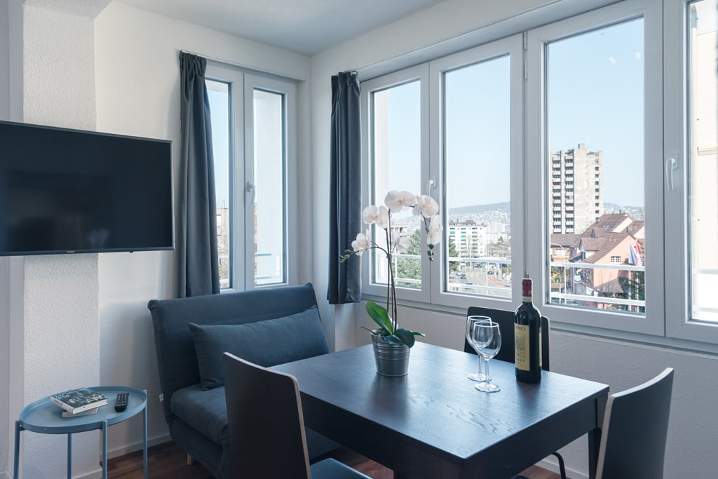 Spacious living room with large windows offering a city view, dark blue sofa, dining table with chairs, TV, and decorative orchid plant