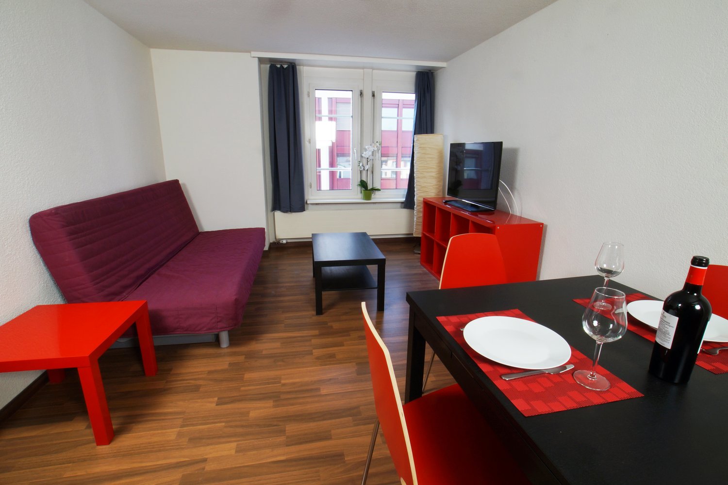 Spacious living room with hardwood flooring, large windows, a burgundy sofa, a black coffee table, a red TV stand, and a dining table with red accents. The room has a modern and minimalist design with pops of color.
