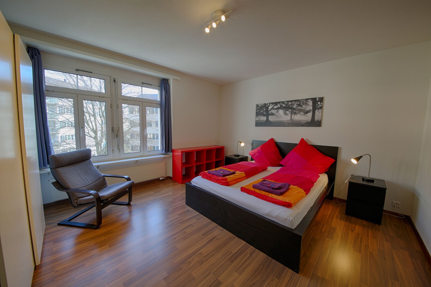 Spacious bedroom with large windows, hardwood floors, black bed frame, red and orange bedding, red storage unit, and a gray armchair