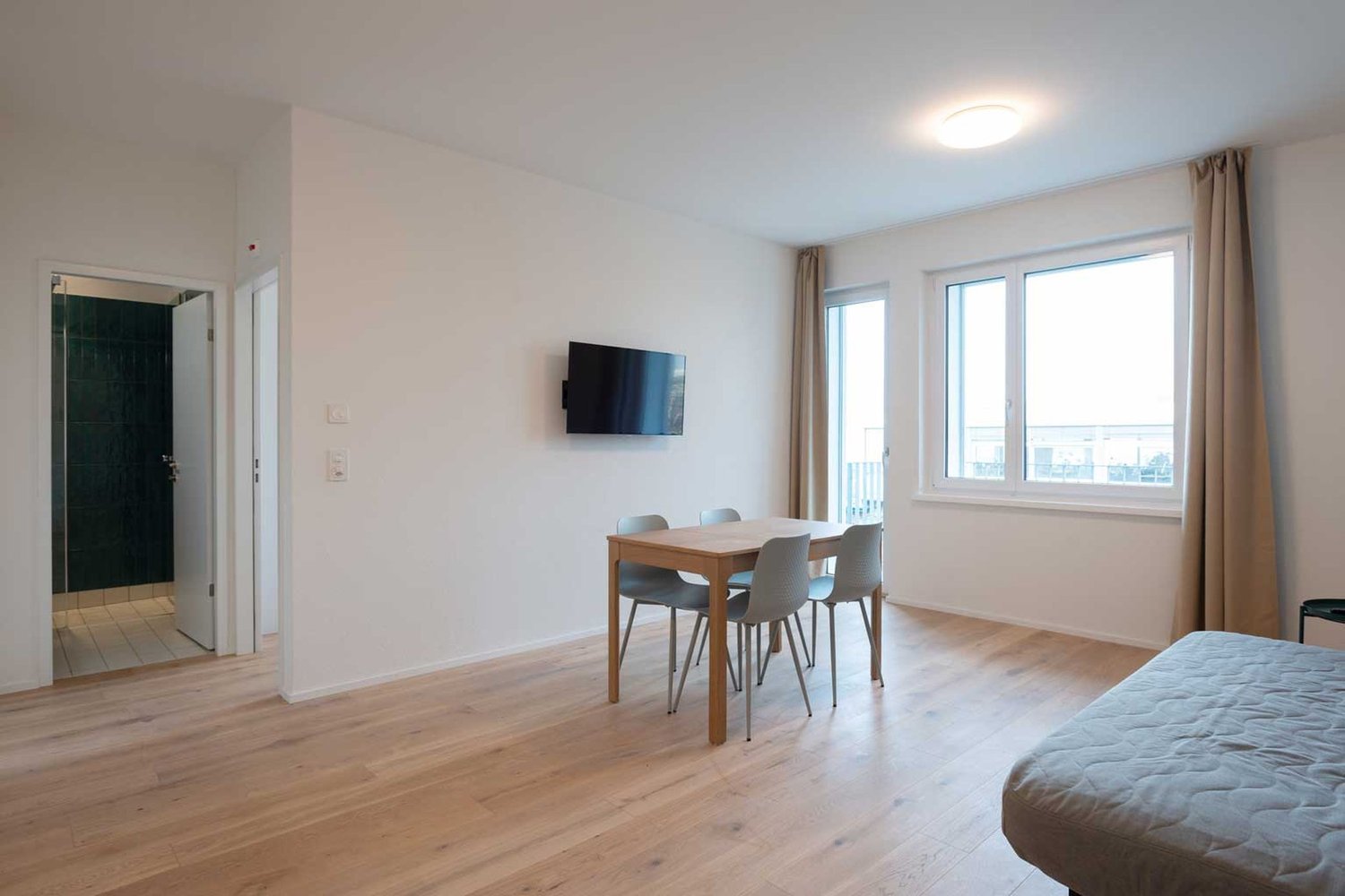 Spacious studio apartment with hardwood floors, white walls, large windows, dining table with 4 chairs, and a TV mounted on the wall. The apartment has access to a balcony or terrace.