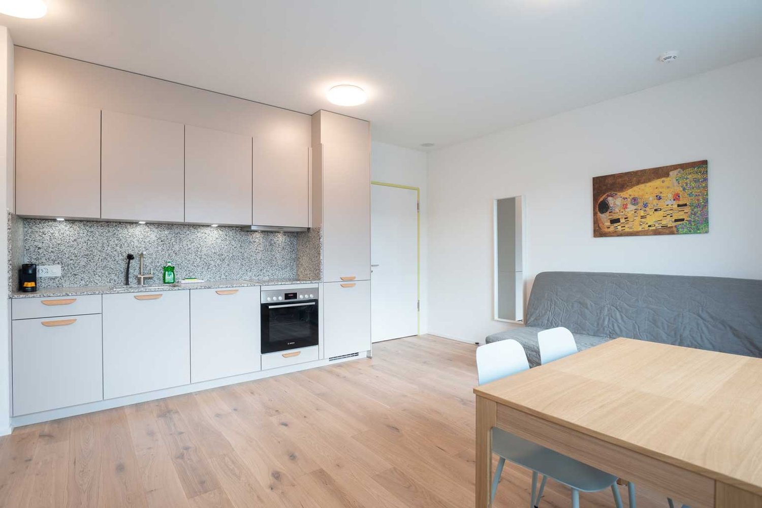 The image shows an open-plan kitchen and living room area. The kitchen features light-colored cabinets, a granite or stone backsplash, and a built-in oven and dishwasher. The living room area has a gray sofa and a wooden dining table with chairs. The floo