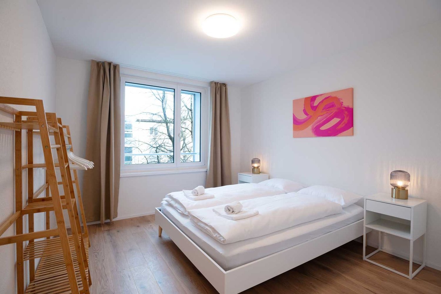 Spacious bedroom with white walls, large windows, hardwood flooring, a white bed with white bedding, two bedside tables with lamps, and a colorful abstract painting on the wall. The room also has a wooden ladder-like structure, possibly for storage or dec