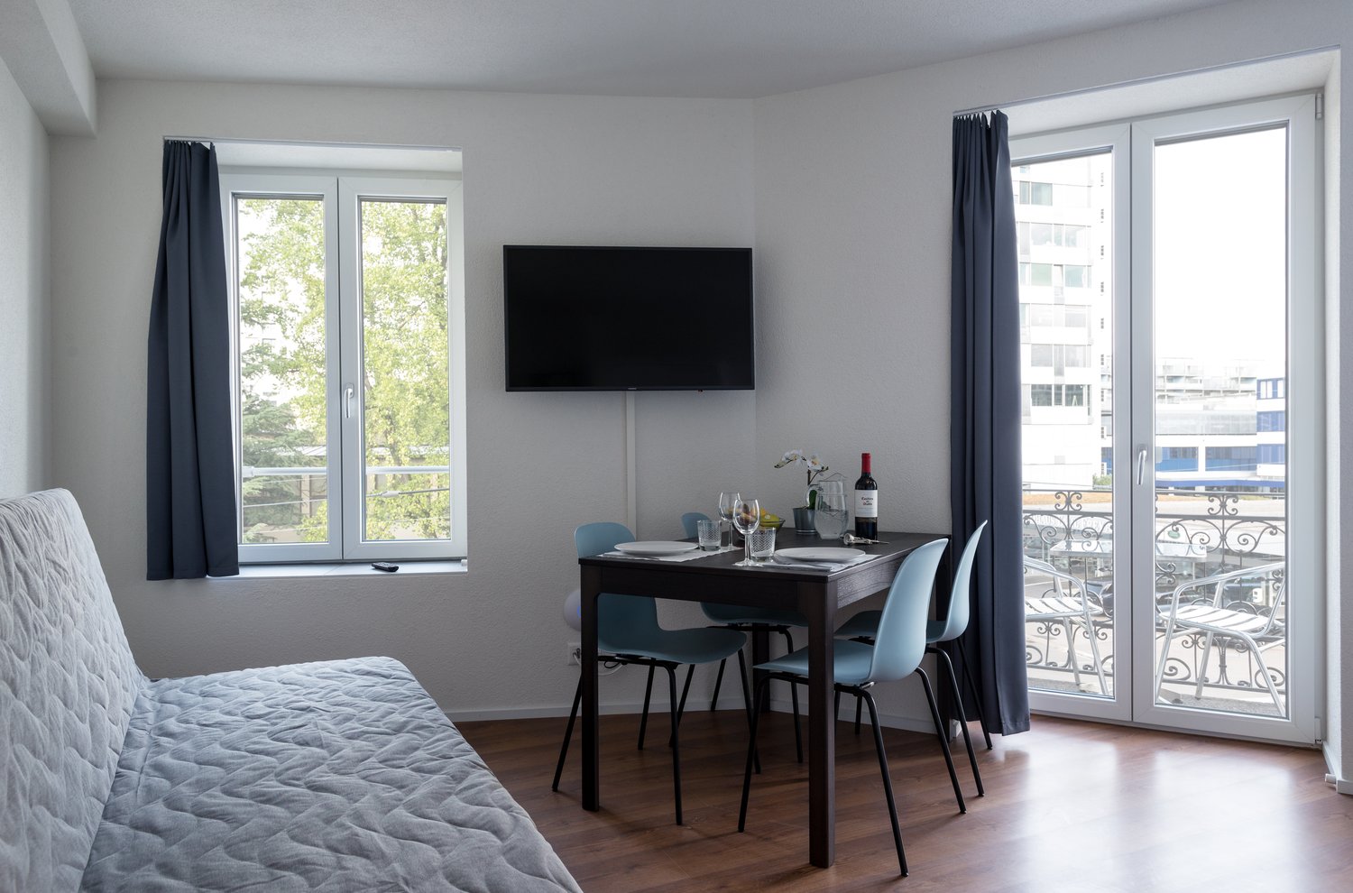 The image shows a studio apartment with a living/sleeping area, a dining area, and a balcony. The living area has a large window with a view of the outdoors, a gray quilted bedspread, and a mounted TV. The dining area has a dark wooden table with blue cha