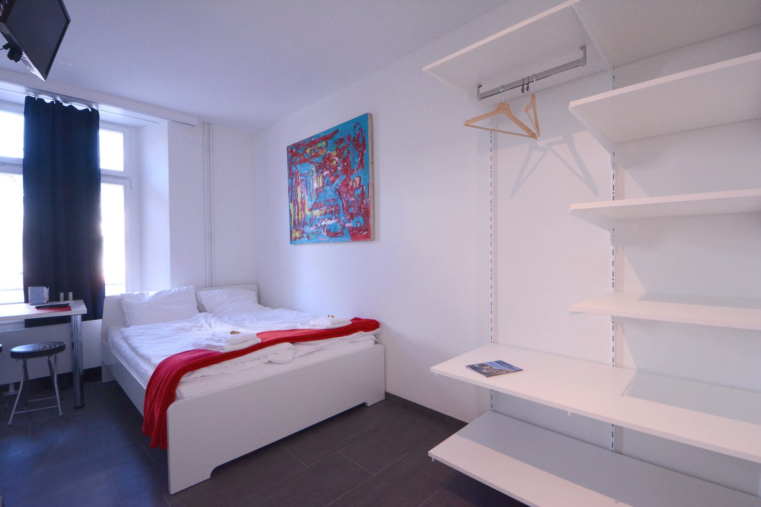 Bright and spacious bedroom with white walls, a large bed, a colorful abstract painting on the wall, and built-in shelving units. The room has a modern and minimalist design.