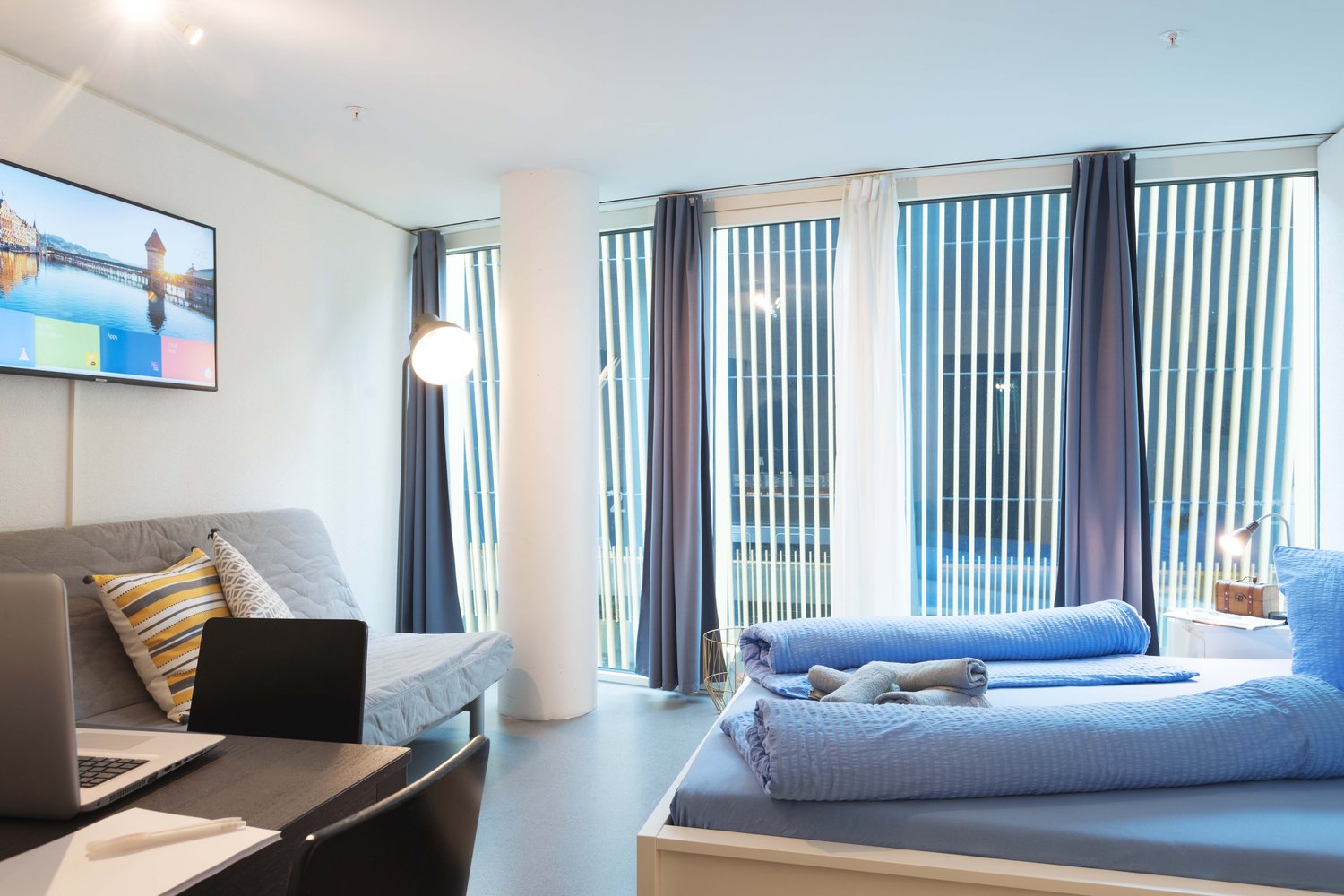 Spacious bedroom with large windows, gray sofa, laptop on a table, and a bed with blue bedding
