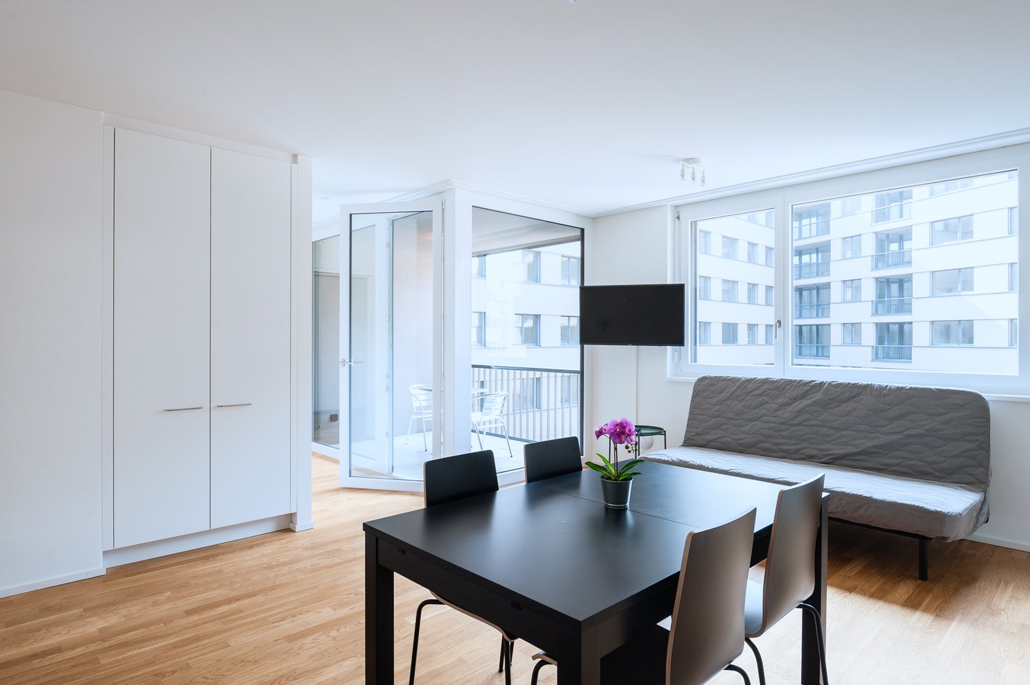 Spacious open-plan living area with large windows, hardwood floors, black dining table and chairs, gray sofa, built-in wardrobe, and access to a balcony or terrace with a view of the surrounding buildings.